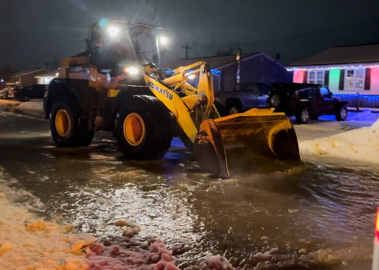 A loader pushes water down a street with houses on either side of it. 