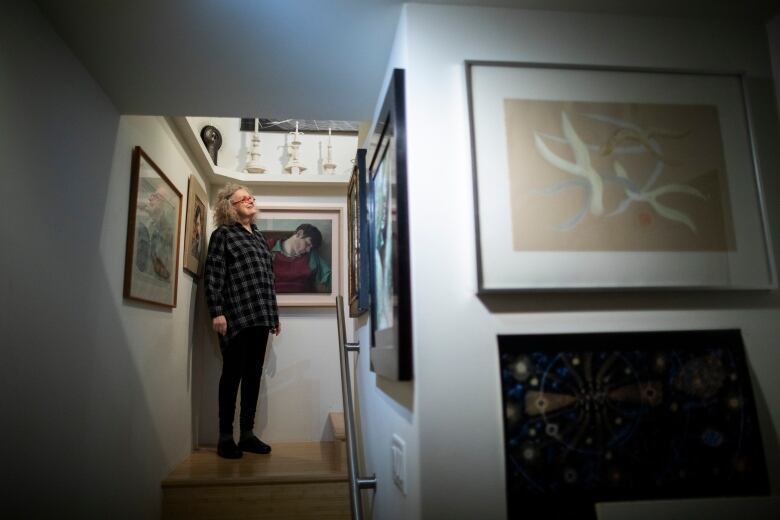 Artist Susanna Blunt stands in a stairwell surrounded by portraits she has painted. Paintings can be seen on all the surrounding walls, and surfaces are covered in sculptures.