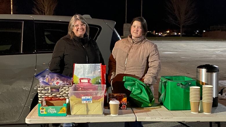 two women in winter coats stand behind folded table in parking lot. table is piled with bags and food.