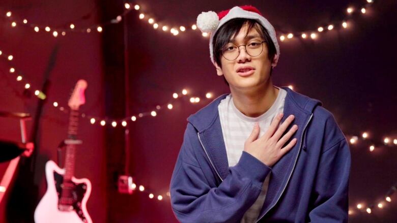 Young man, 18, has dark hair and bangs. He wears a red and white Santa hat and has round, wire-framed glasses. He is wearing a blue hoodie opened with a white T-shirt with light grey, horizontal stripes. He has his left arm up with his hand on his chest. In the background are white holiday lights strung horizontally on the back wall. An electric guitar and drum kit are on the left side of the picture in the background. 