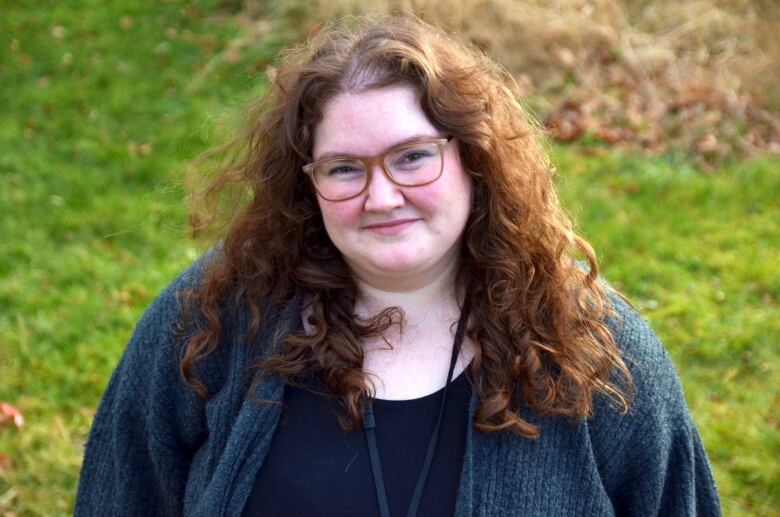 A woman with glasses wearing a grey sweater standing in a field with trees in the background. 