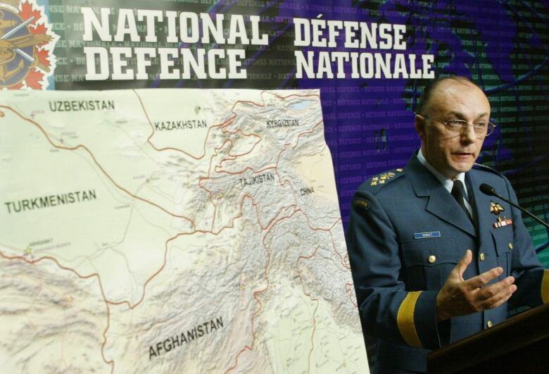 A man with glasses, dressed in a military uniform, speaks to reporters while next to him is a map of Afghanistan. 