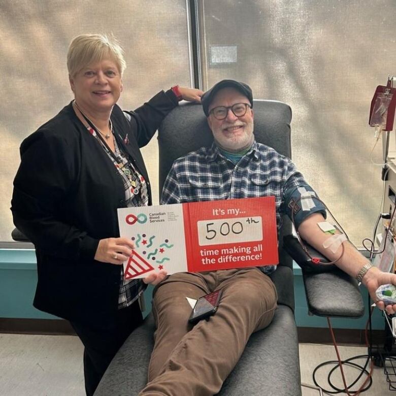 A man sits in a chair as he gives blood. He and a woman are holding a sign that reads 'It's my 500th time making all the difference.'