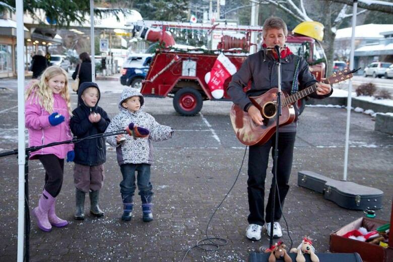 A man plays a guitar outdoors where children dance beside him.