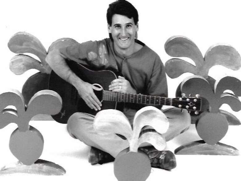 A black-and-white photo of a man with a guitar surrounded by fake radishes.