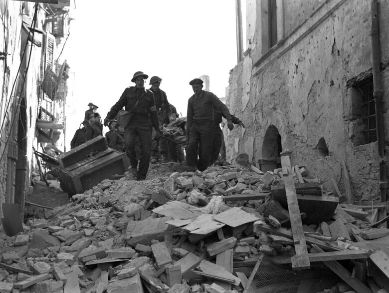 A black and white photo shows a rubble-filled street in Ortona, Italy, as Canadian soldiers carry an injured fellow solider on a stretcher.