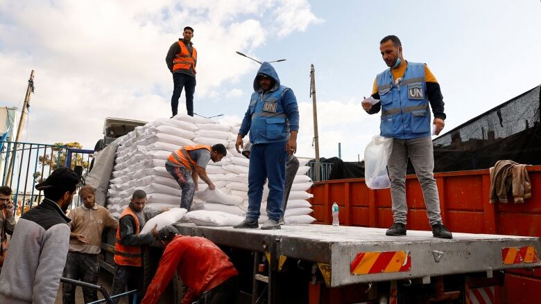 Men wearing blue UN vests or orange safety vests unload bags of flour from the back of a flatbed truck. 