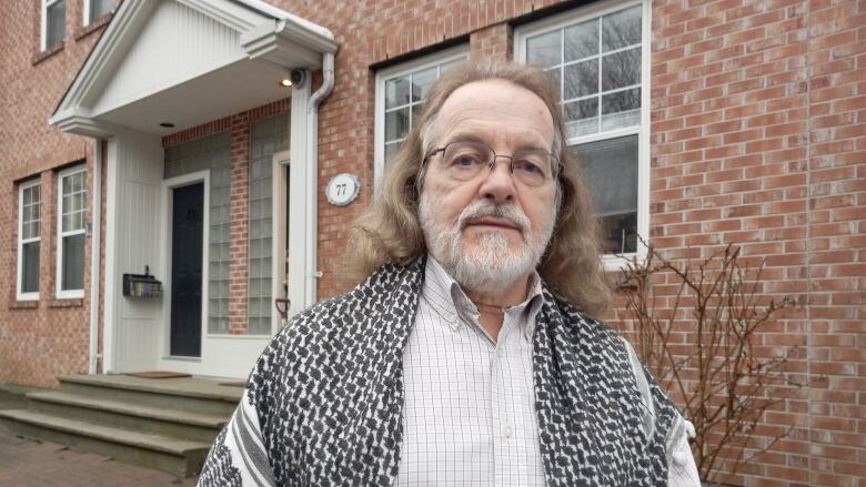 Man with white beard with hair to shoulders in dress shirt and shawl over shoulders standing in front of brick house.