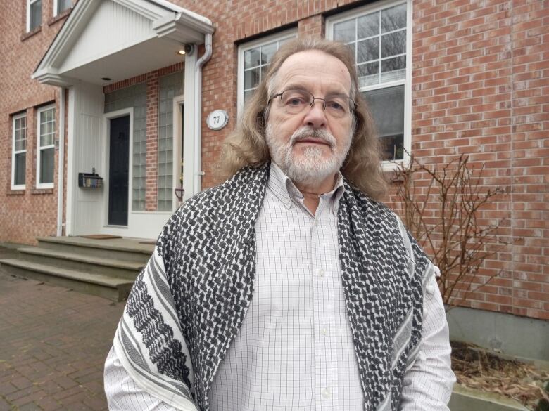 Man with white beard with hair to shoulders in dress shirt and shawl over shoulders standing in front of brick house.