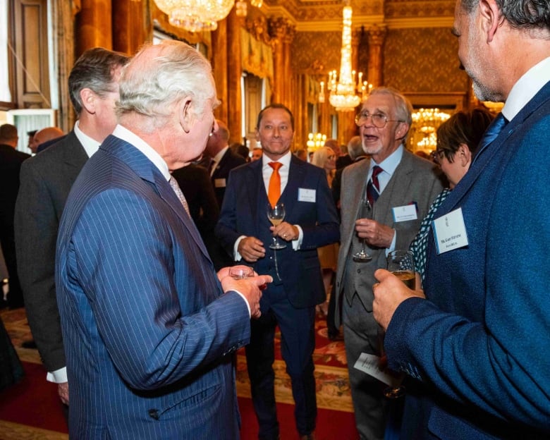 Five people holding wine glasses listening to King Charles.