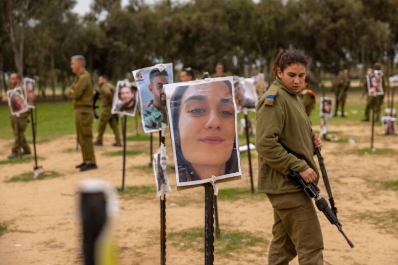 A female solder holding a large gun walks in a field among a display of large photos of faces