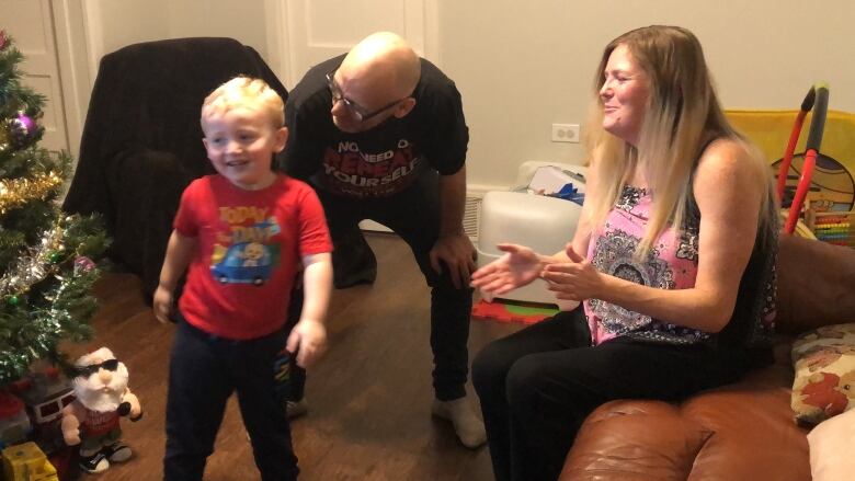 A young boy wearing a red t-shirt and black pants standing next to his bald father and his mother