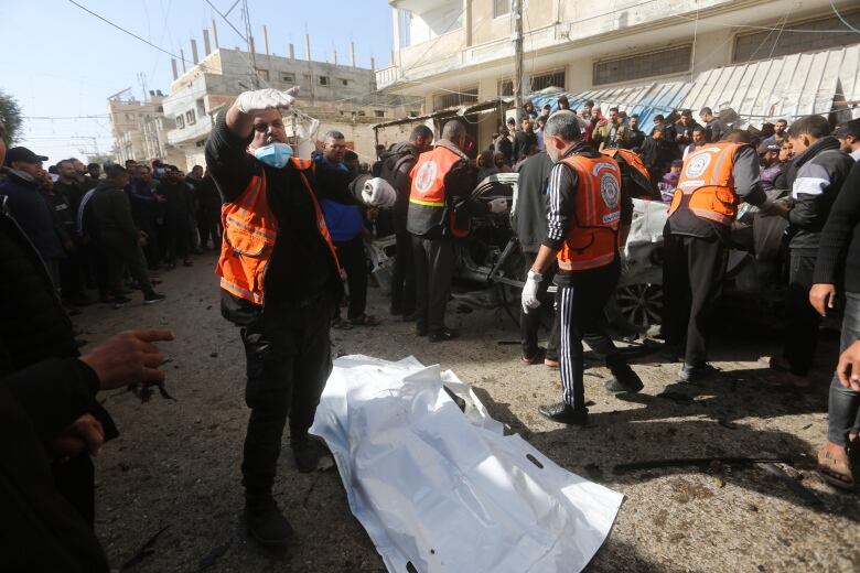 A man in an orange vest stands next to a shrouded corpse.