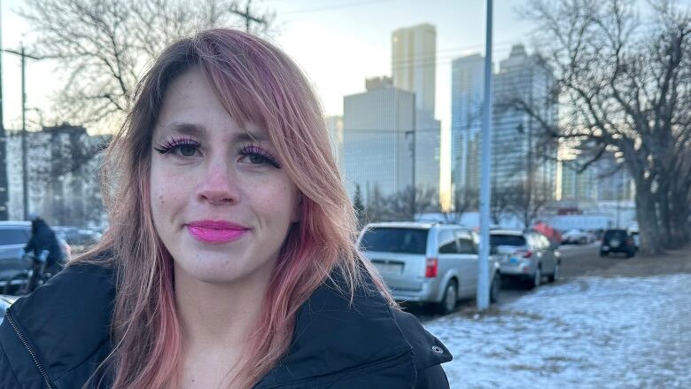 A woman with dyed-pink medium-length hair and long eyelashes smiles at the camera. The woman is wearing a black winter jacket and standing outside. There is snow on the ground. 