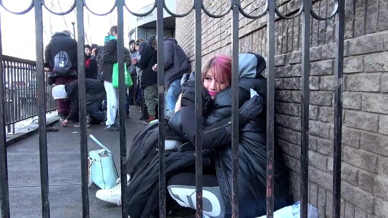 A woman in pink hair wearing a black jackets hugs another person sitting on the ground who's wearing a black puffer jacket. 