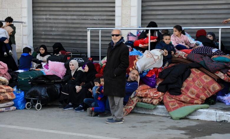 A man wearing sunglasses stands with his hands in his pockets as men, women and children sit on piles of belongings on the street behind him.