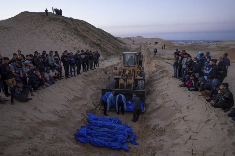 Bodies in blue bags lie in a trench near a bulldozer, with men gathered on either side of the trench.