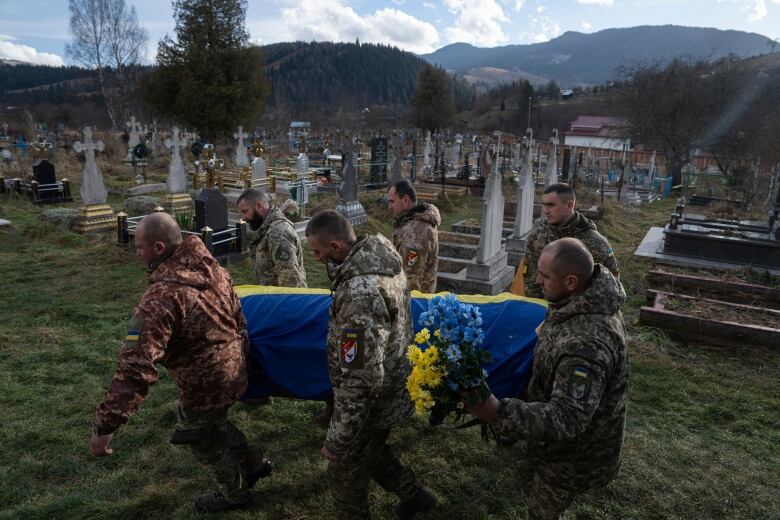 Six men in camoflauge carry a coffin draped in a blue and yellow flag. One of the men carries blue and yellow flowers in his other hand.