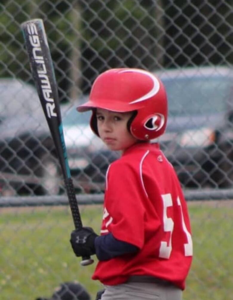 A young man in a red baseball uniform and helmet holds a black baseball bat.