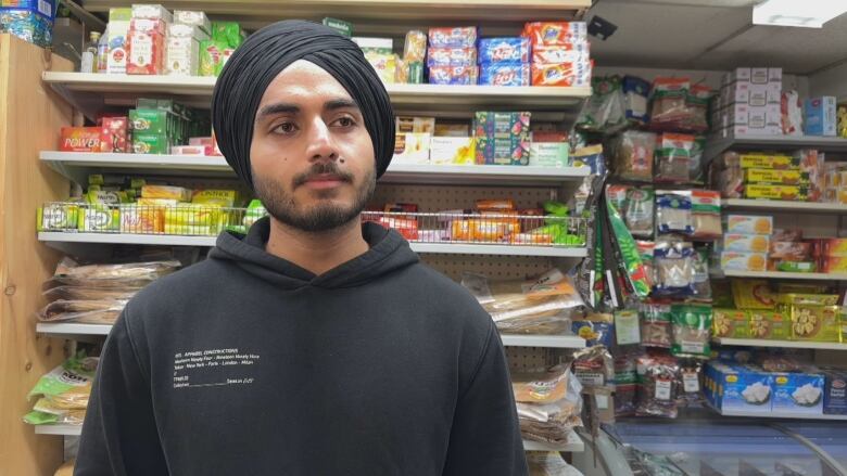 A man wearing a black turban and hoodie is shown looking at the camera with Indian snacks and spices on the store shelves behind him. 