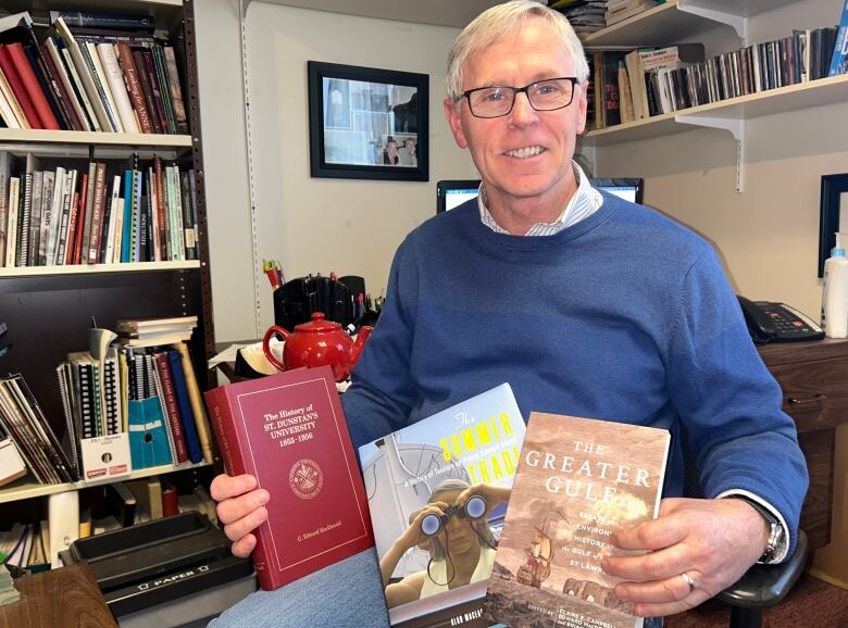 A man in a blue sweater sits in an office holding three books.