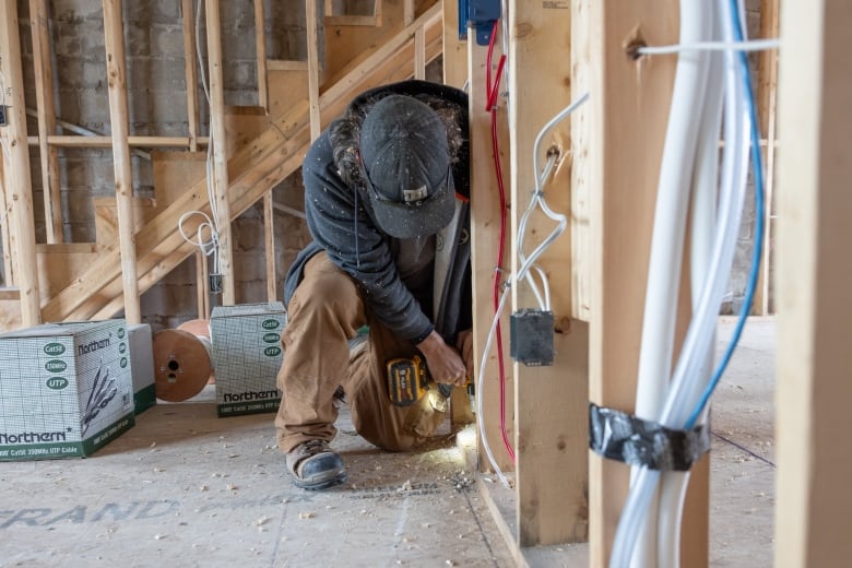 A construction worker is crouched down inside of a structure that looks incomplete. They are holding a drill and are in the process of using it.