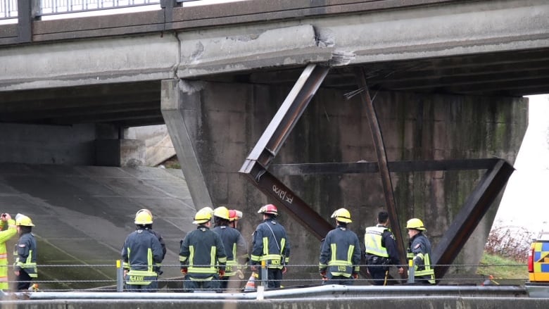 A group of firefighters stands below an overpass damaged by a collision with a semi-truck.