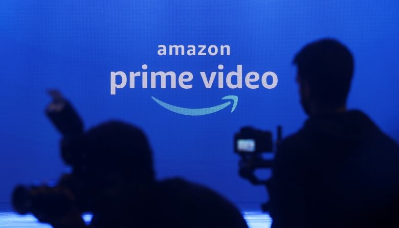 Two cameramen are silhouetted in front of a blue Amazon Prime Video logo.