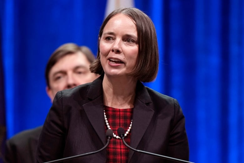 A woman with short, dark hair wearing a black blazer over a tartan blouse with pearls speaks at a microphone in front of a blue curtain as a man looks on. 