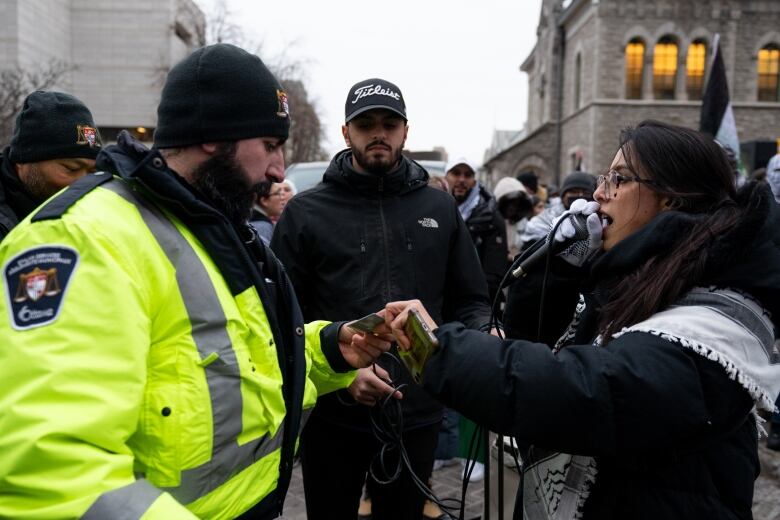 A protester speaks into a microphone as a bylaw officer checks an ID card, or something similar.