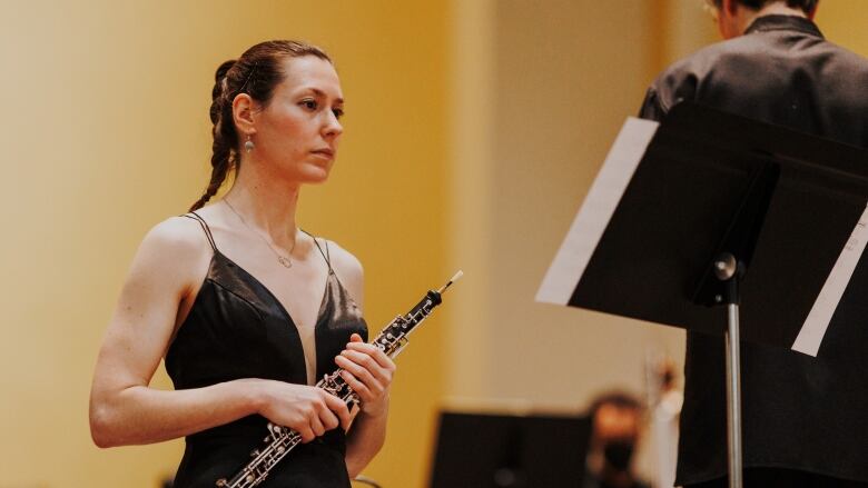 A woman in a black dress looks at sheet music while holding an oboe. 