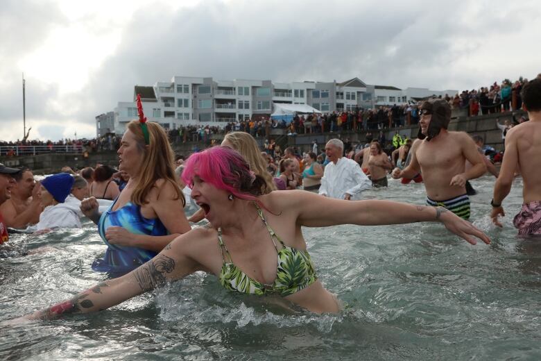 Dozens of people mill around in waist-deep water while a row of people behind them watch.