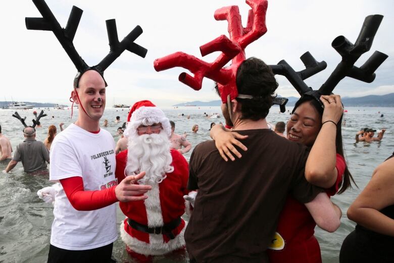 Three people wearing costume antlers, and one person wearing a Santa costume, pose at a beach while others are behind them in the water.