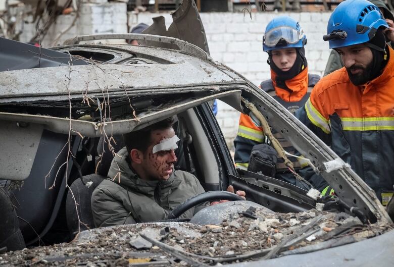 A person with facial wounds and bandages sit inside a destroyed car as rescuers in high-viz gear outside the vehicle talk to them.