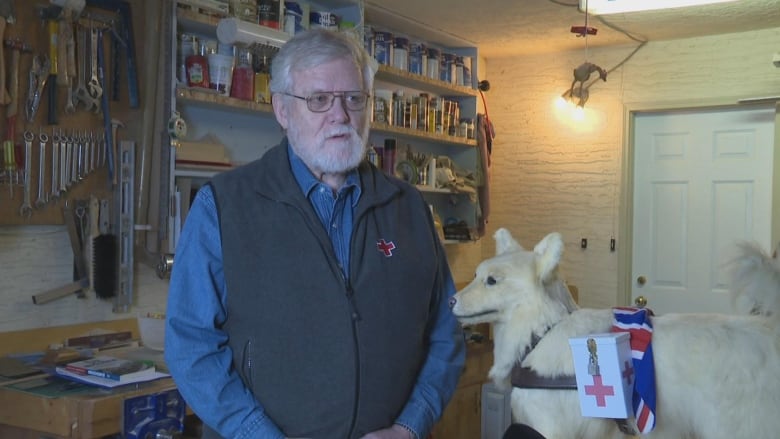 A man wearing a Red Cross sweater-vest stands next to a taxidermied white dog in a garage.