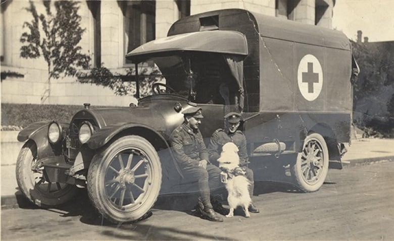 Two soldiers pet a dog while seated on the step of an ambulance in a black-and-white picture.