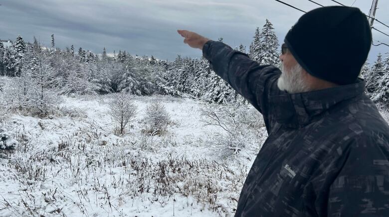 Man in winter coat and hat points at land with snowy trees.