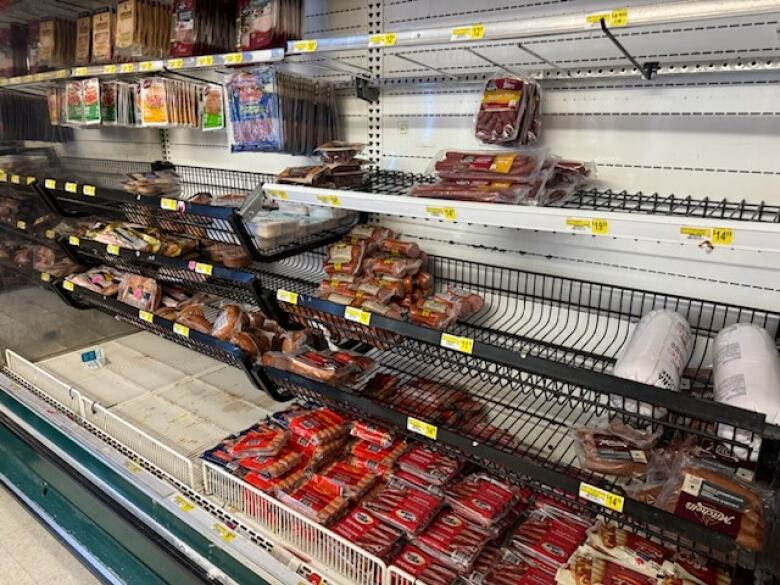 A half-stock meat shelf at a grocery is shown. Some products like packaged hot dogs can be seen while other sections are empty.