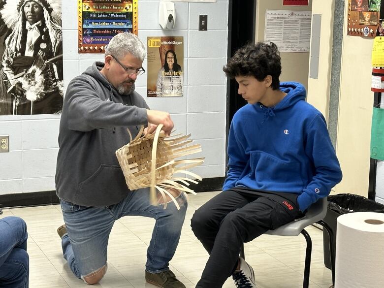 A man with grey hair wearing a grey hoodie and jeans bends on one knee next to a teen boy wearing a blue sweater. The man holds up a half-finished basket.