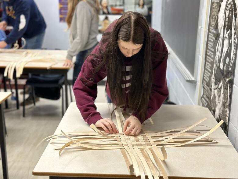 A teenager with long dark hair bends over a table, weaving strands of wood into a basket.