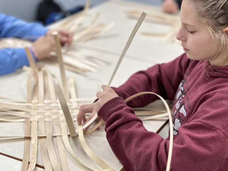 A teenager wearing a red sweater sits at a table weaving strands of wood into a basket.