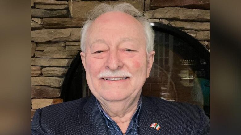 A man with white hair in a blue jacket and shirt smiles for the camera.