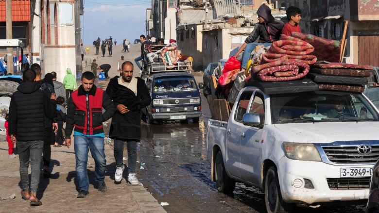 People trudge along the side of a road while trucks, loaded down with personal belongings, drive by.