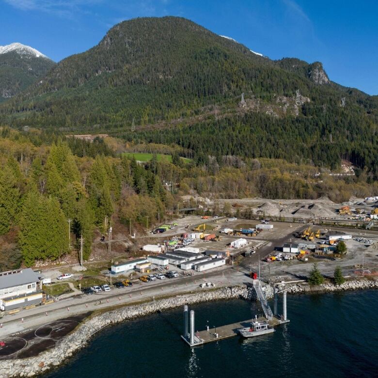 An aerial photo shows a construction site along the water with mountains in the back. 