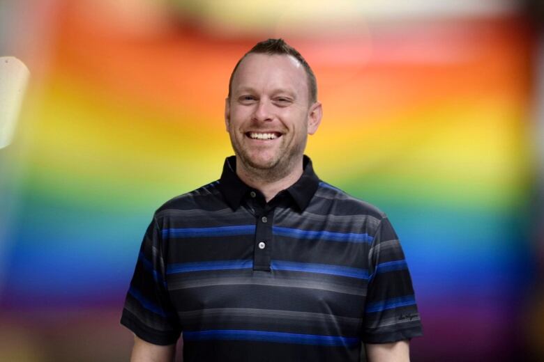 A man smiles with a rainbow background behind.