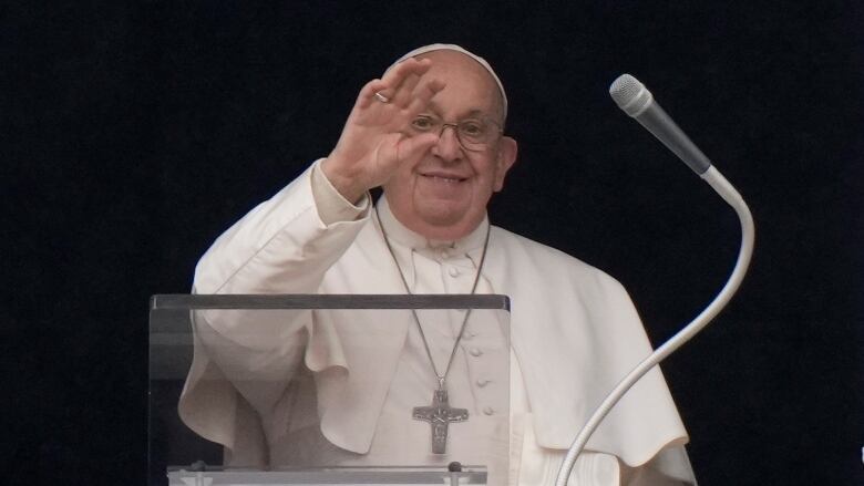 The bespectacled pope waves a hand to an unseen crowd, while wearing a large cross around his neck.