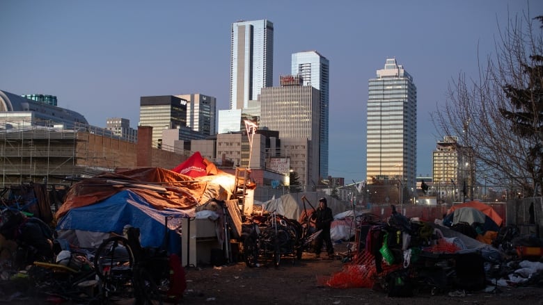 A man stand in the middle of a cluster of tents. The downtown Edmonton skyline can be seen in the background. 