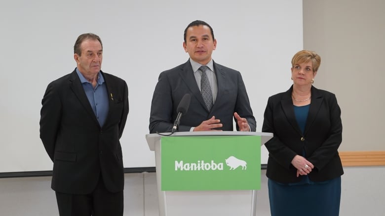 A man speaks at a microphone, with a man on one side of him and a woman on the other.