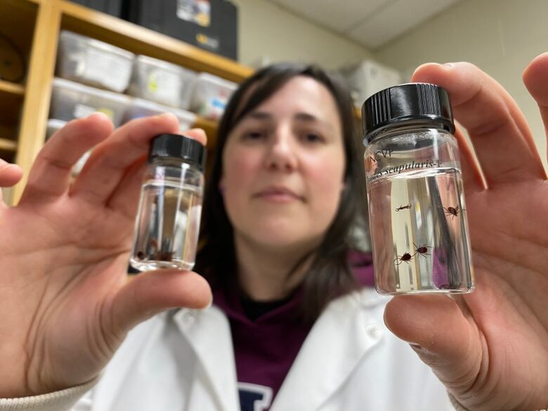 A woman can be seen wearing a lab coat, she is holding two vials with deer ticks in them. 