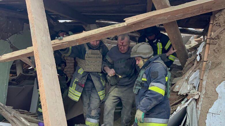 An older man with blood on his face is held by a man in a rescue uniform and helmet near a building with fallen and damaged pieces of wood.